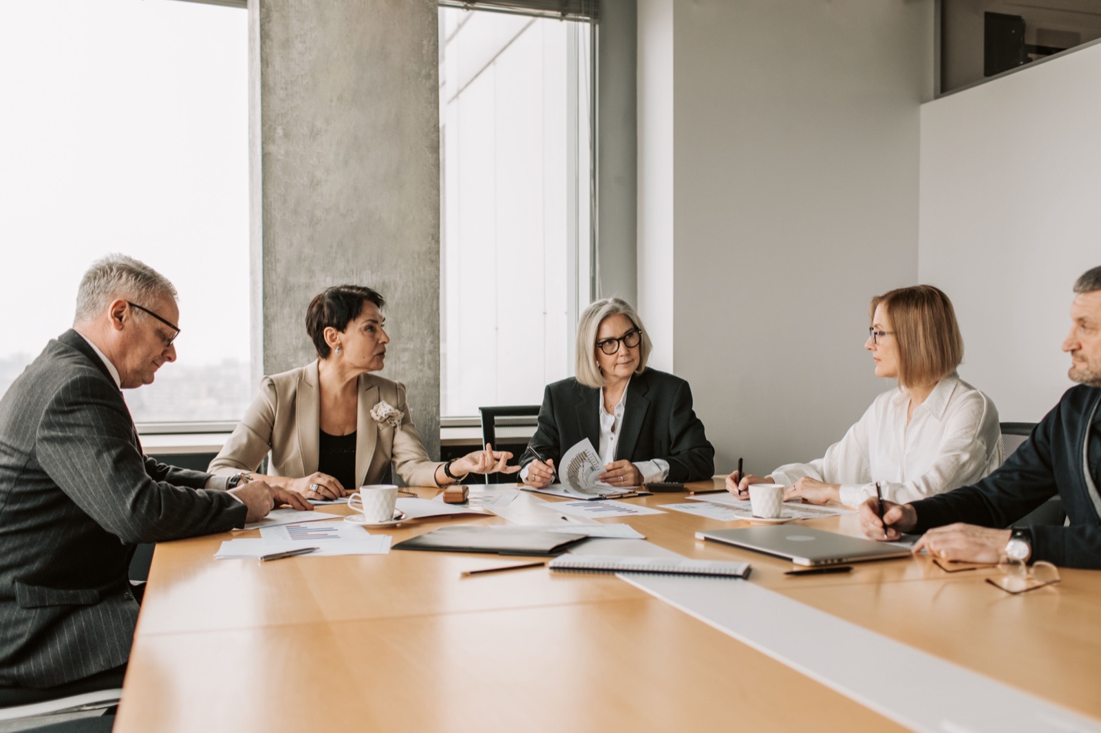 Five board members around a meeting table with papers, listening as one of them speaks
